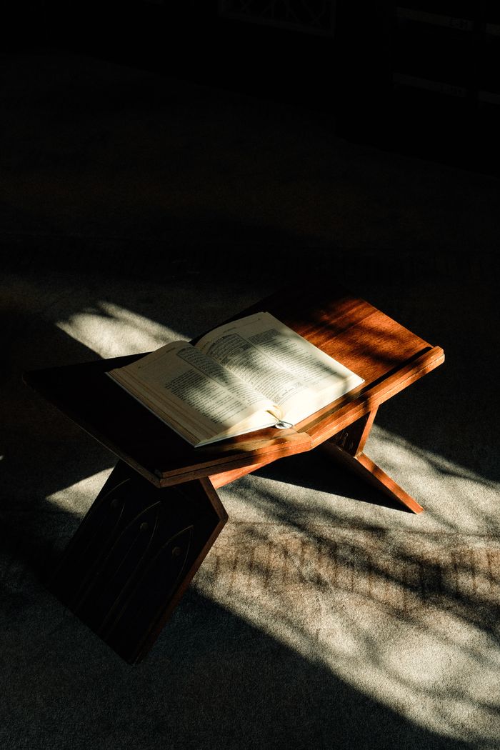 An open Quran rests on a wooden stand illuminated by sunlight, casting shadows across the pages.