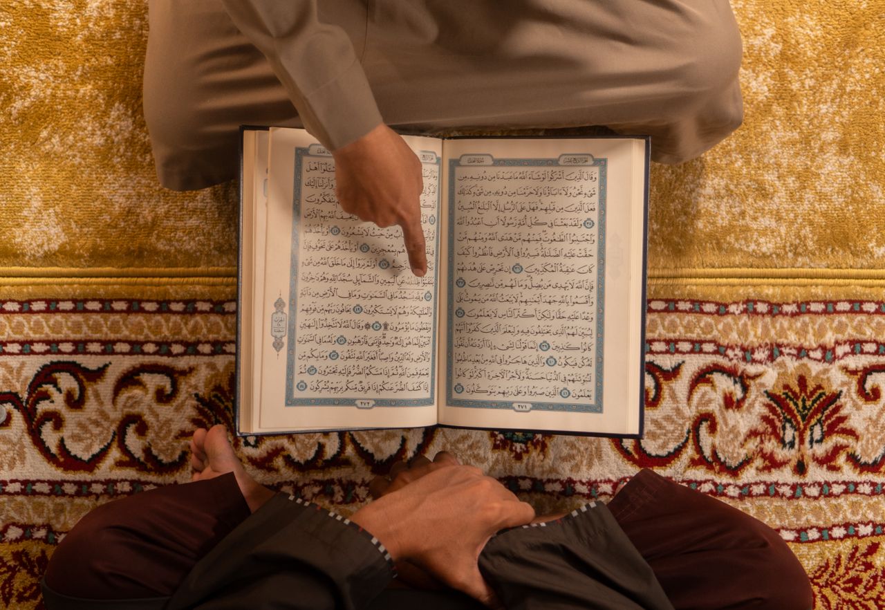 A person pointing to an open Quran while sitting on a prayer rug, emphasizing Islamic teachings.